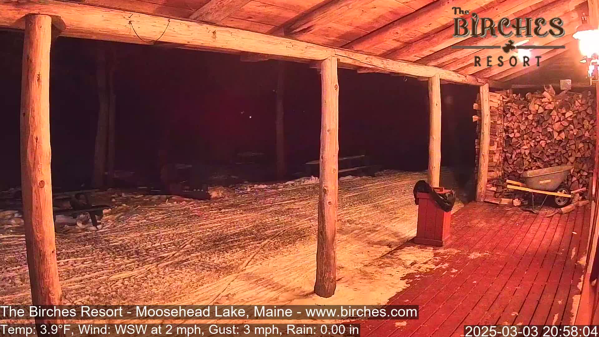 A snow-covered outdoor patio with wooden posts and a red wooden floor is illuminated at night under a dark sky.