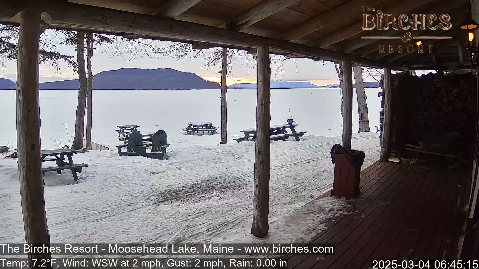 A snow-covered lakeside scene is viewed from a wooden porch, showing picnic tables and Adirondack chairs in the distance with mountains in the background under a twilight sky.