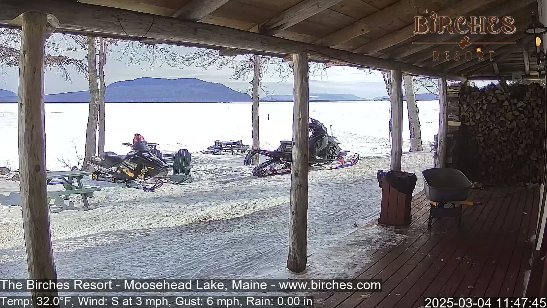 From a wooden porch overlooking a snow-covered lake and distant mountains, two snowmobiles are parked near picnic tables on a partly sunny day with a south wind.