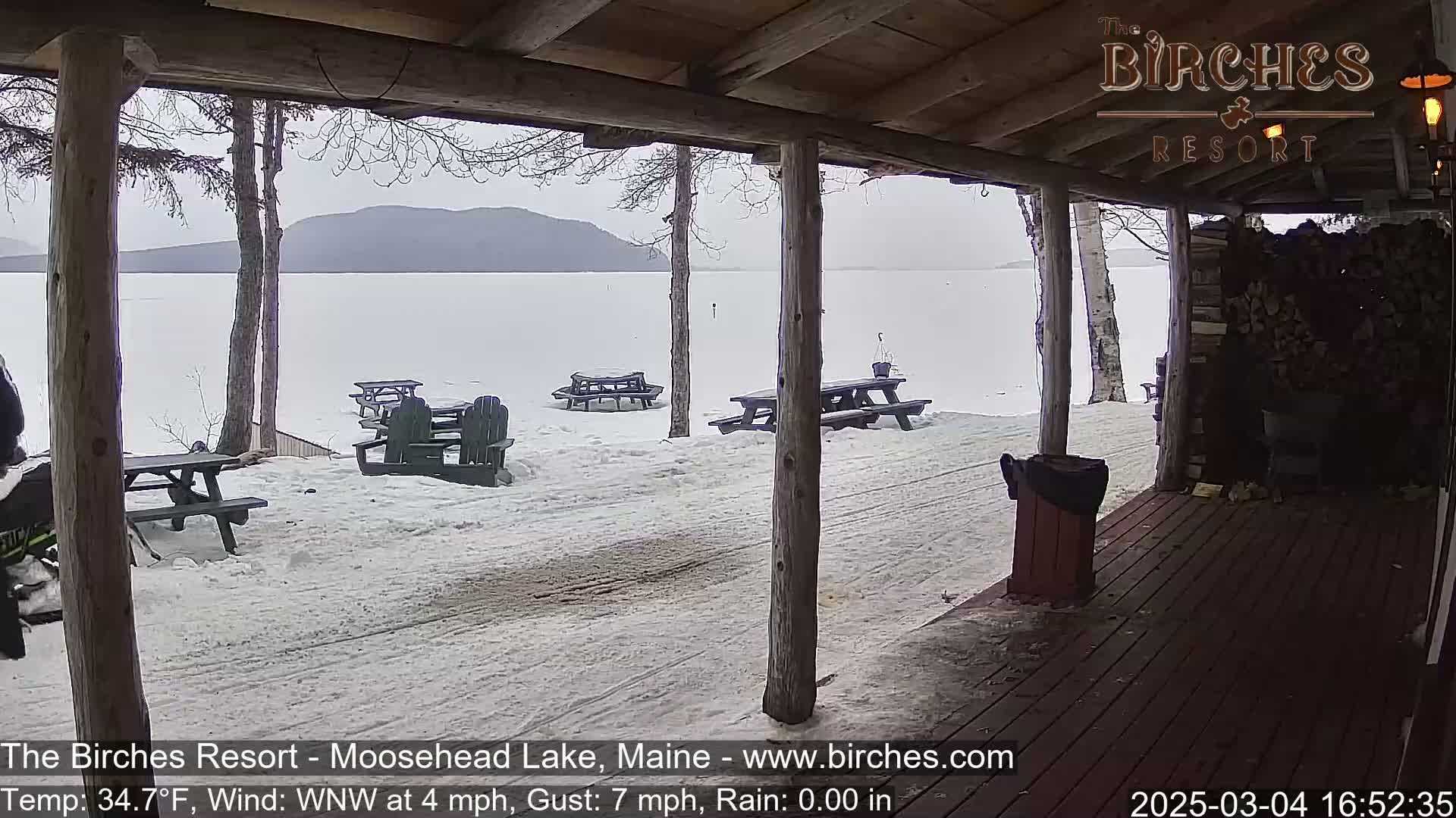 A snow-covered lakeside porch with picnic tables and Adirondack chairs overlooks a frozen lake and distant mountains under an overcast sky.