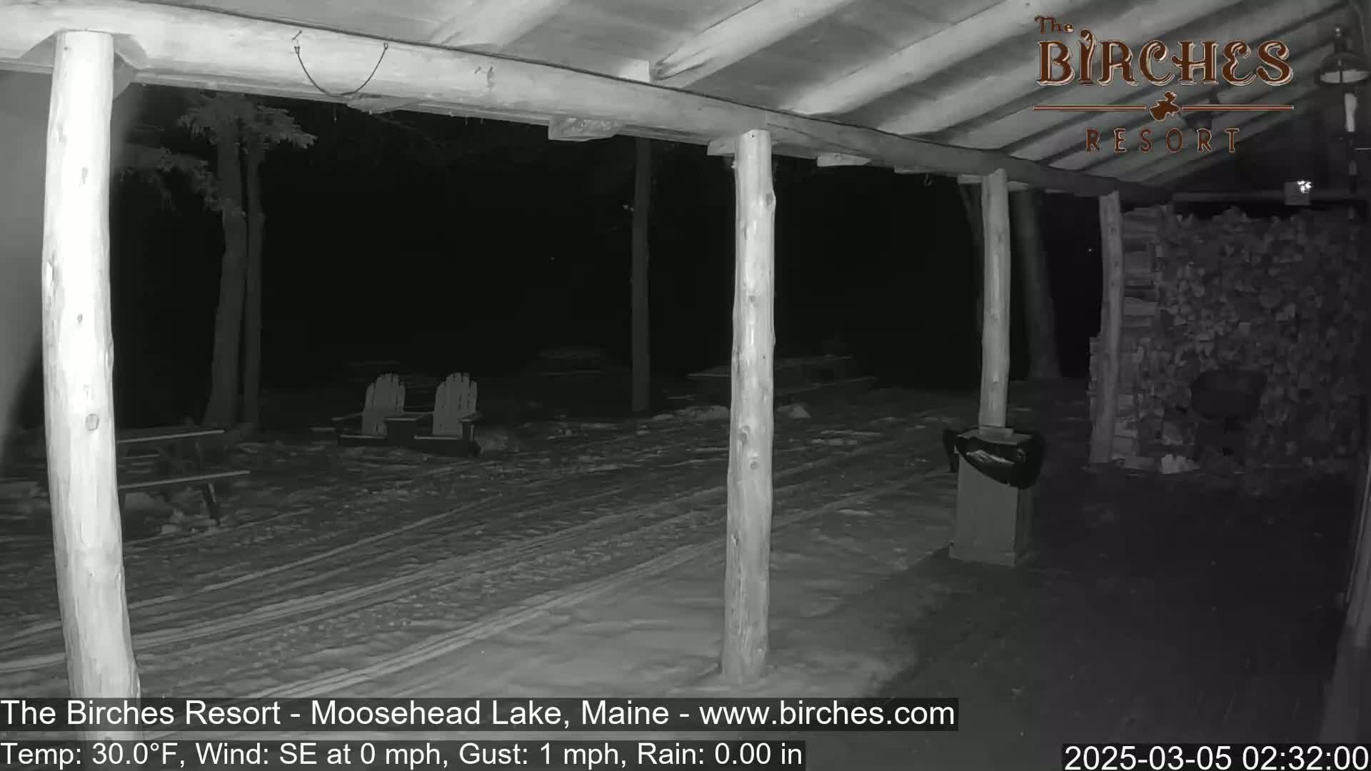 A snow-covered porch at night, viewed from under a covered structure, shows several empty Adirondack chairs and a woodpile in the background during rainy weather.