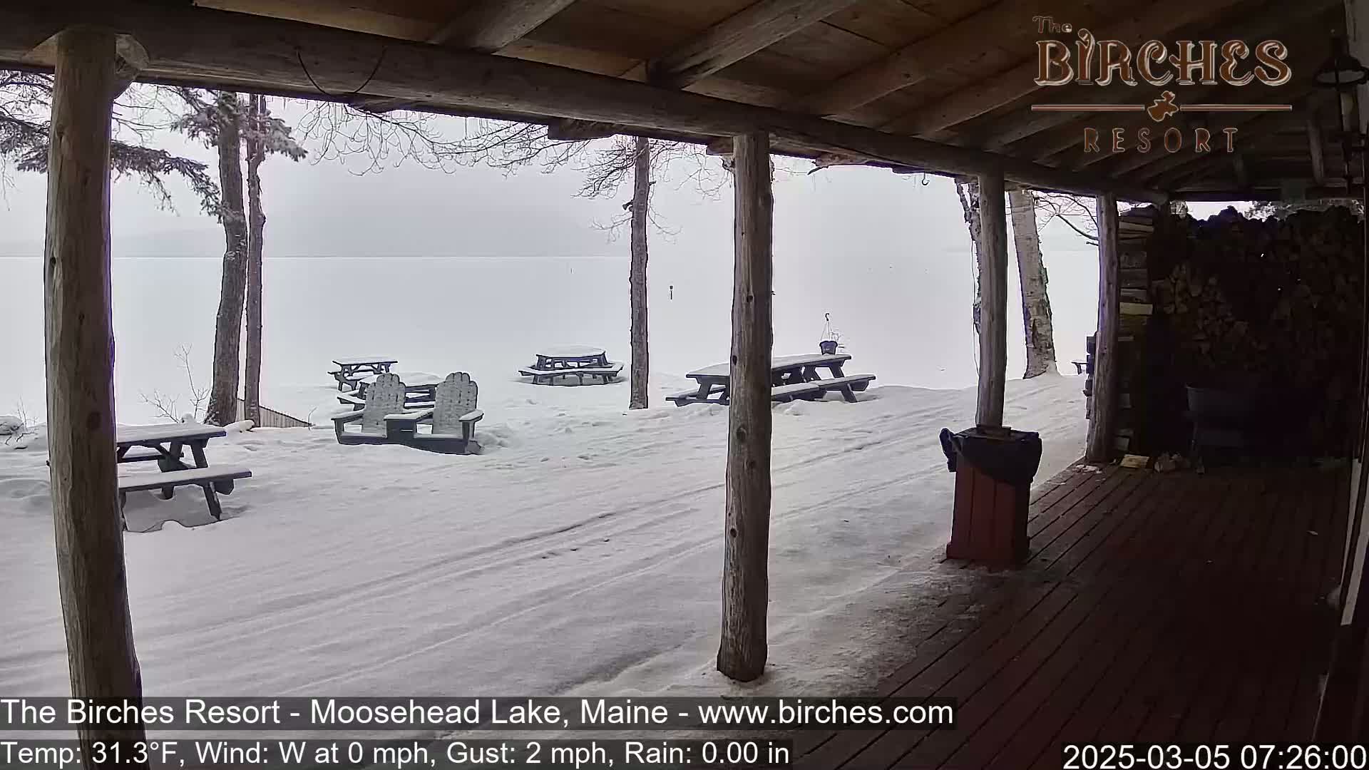 A snow-covered lakeside patio with picnic tables and Adirondack chairs is viewed from a wooden porch under an overcast sky.
