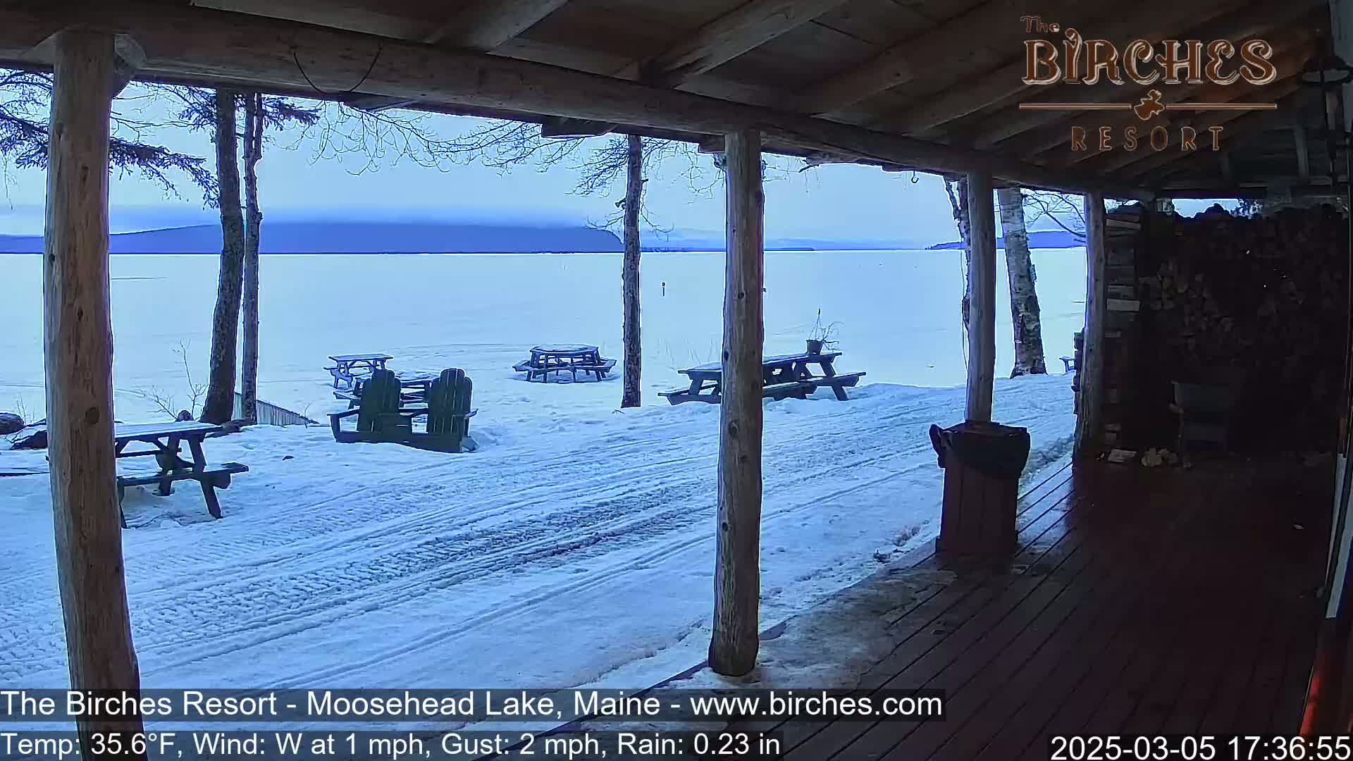A snow-covered lakeside patio with picnic tables and chairs is viewed from a covered porch under a cloudy sky with light rain.