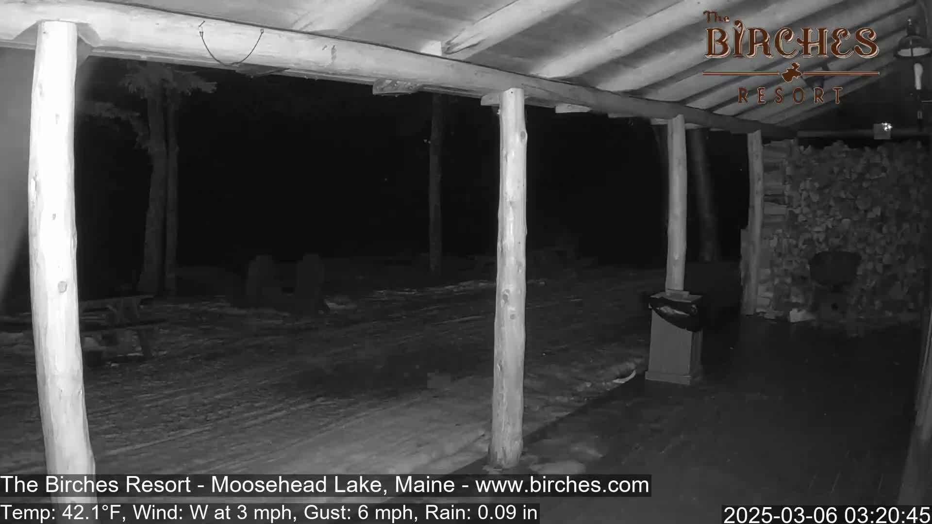 A nighttime view of a covered porch with wooden posts, a woodpile, and light rain falling on snow-covered ground.