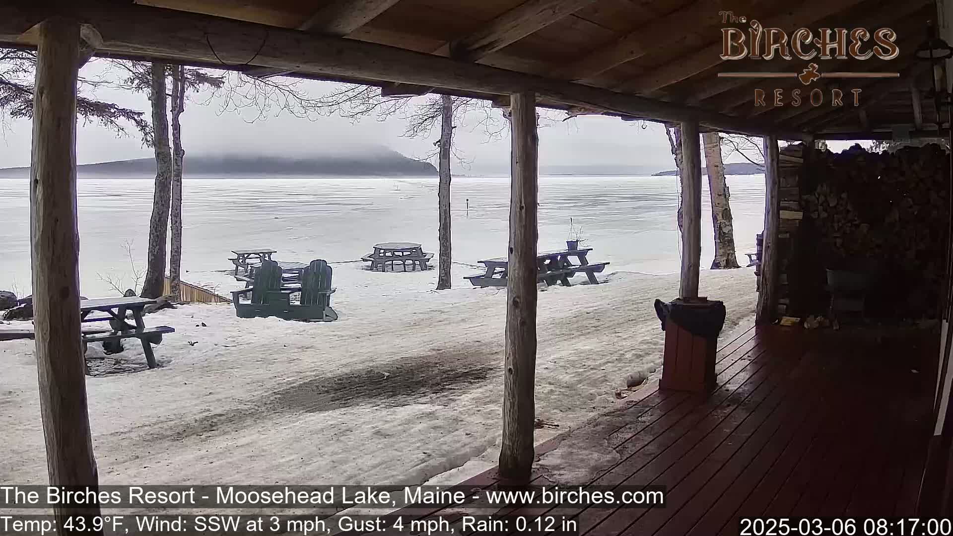 A partially frozen lake is visible from a covered porch, with several picnic tables and chairs scattered on the snow-covered ground in rainy, windy conditions.