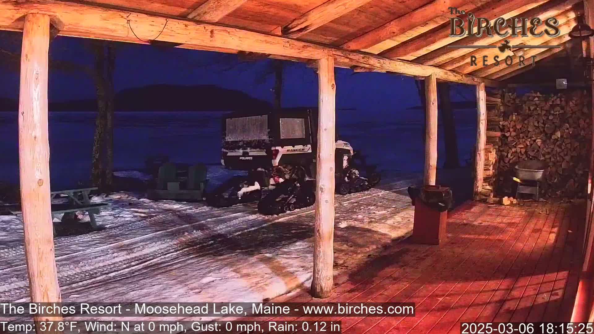 A snow-covered porch with a wooden deck overlooks a dark lake under a nighttime sky, with a Polaris Ranger parked nearby in light rain.