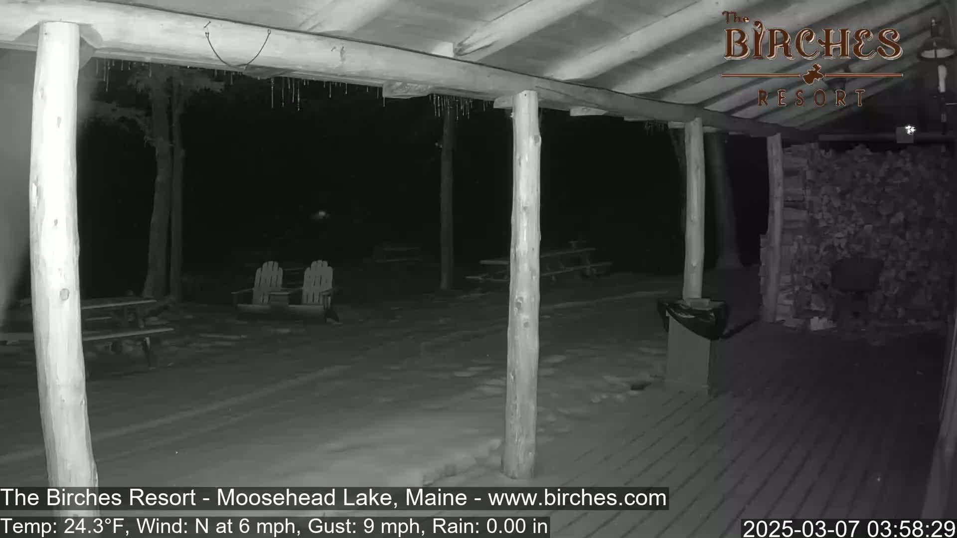 A snow-covered outdoor patio area at night, viewed from under a covered porch with icicles hanging from the roof, shows picnic tables and chairs.