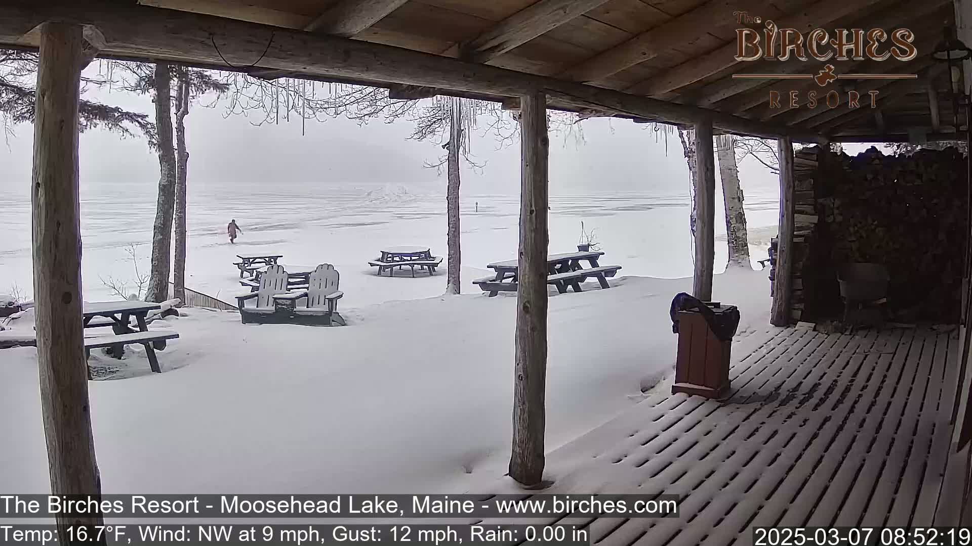 A snow-covered lakeside porch with picnic tables and a person walking on the frozen lake in the background, during a light snowfall with a northwest wind.