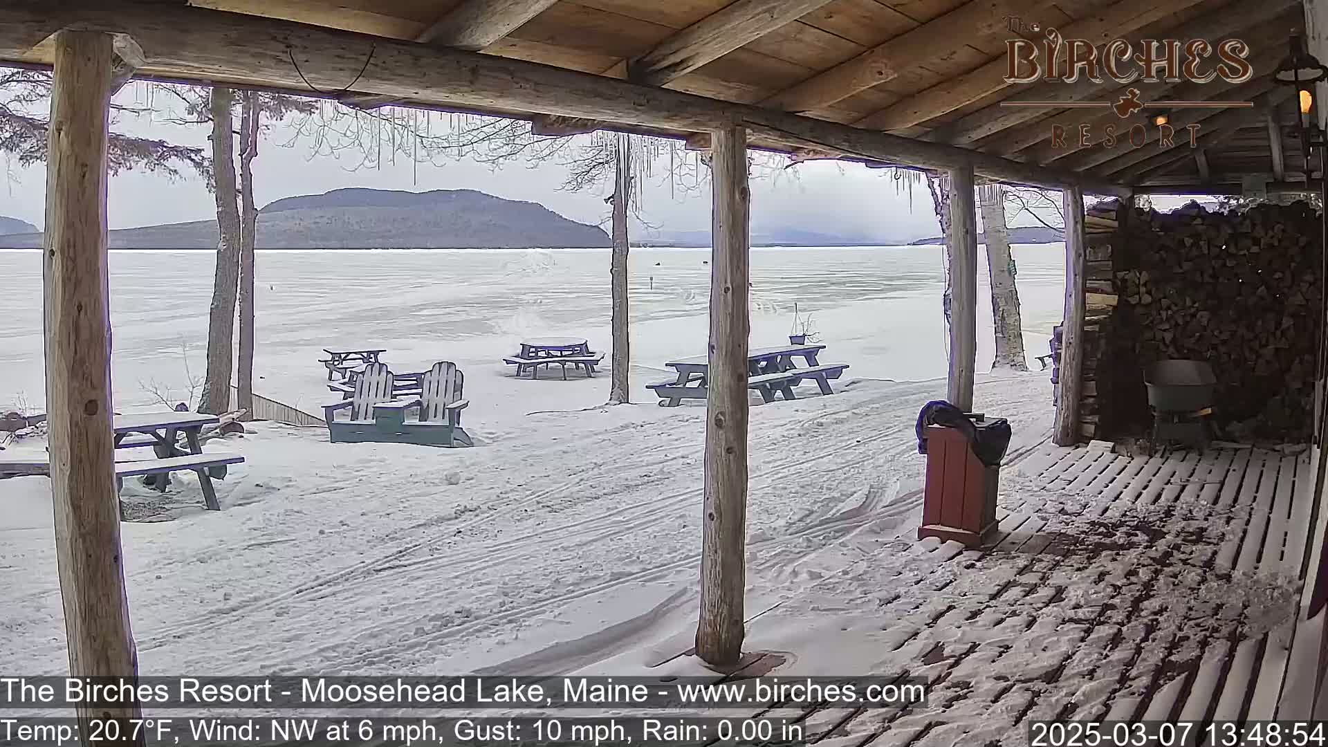 A snow-covered porch overlooks a frozen lake and distant mountains under a cloudy sky.