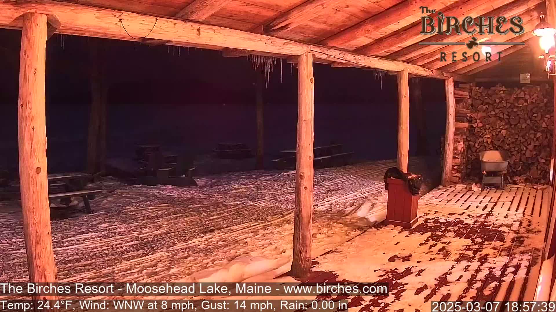 A snow-covered outdoor patio area with picnic tables is seen at night under a wooden covered structure with icicles hanging from the beams.