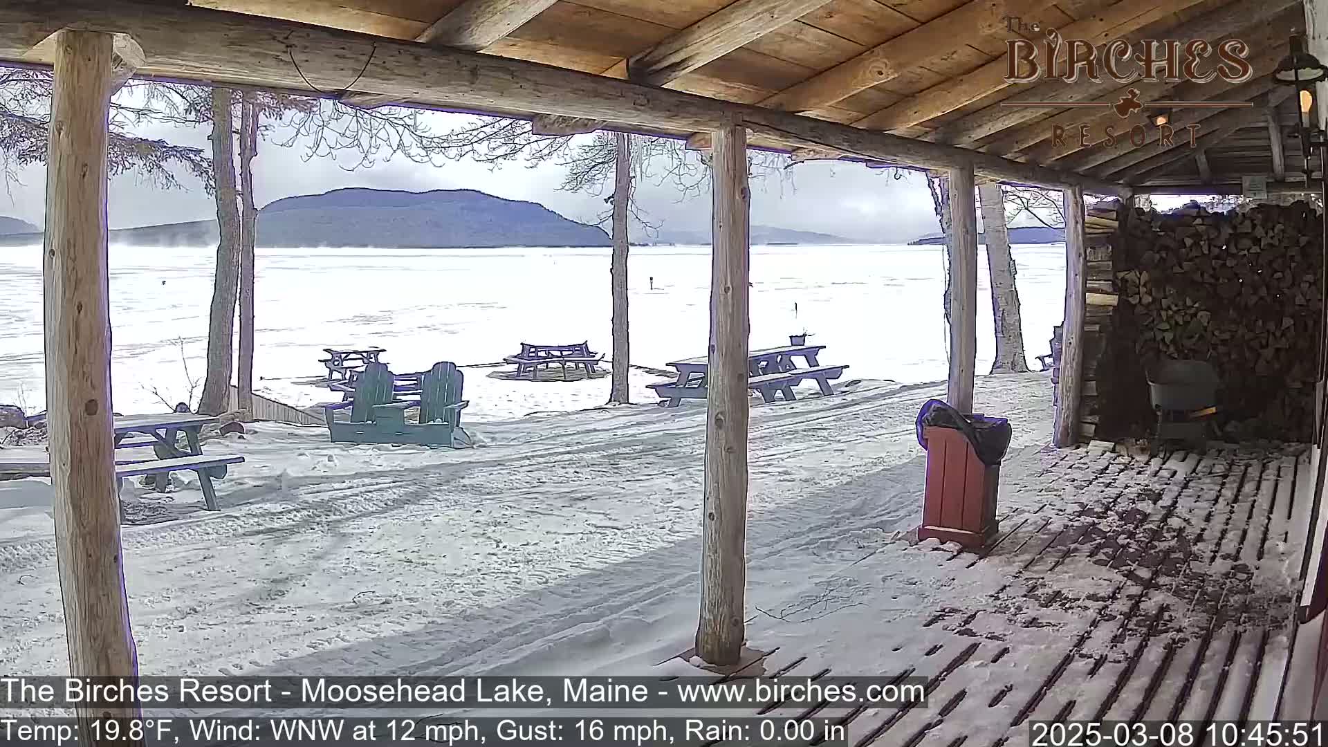 A snow-covered porch overlooks a partially frozen lake with mountains in the background under a cloudy sky.