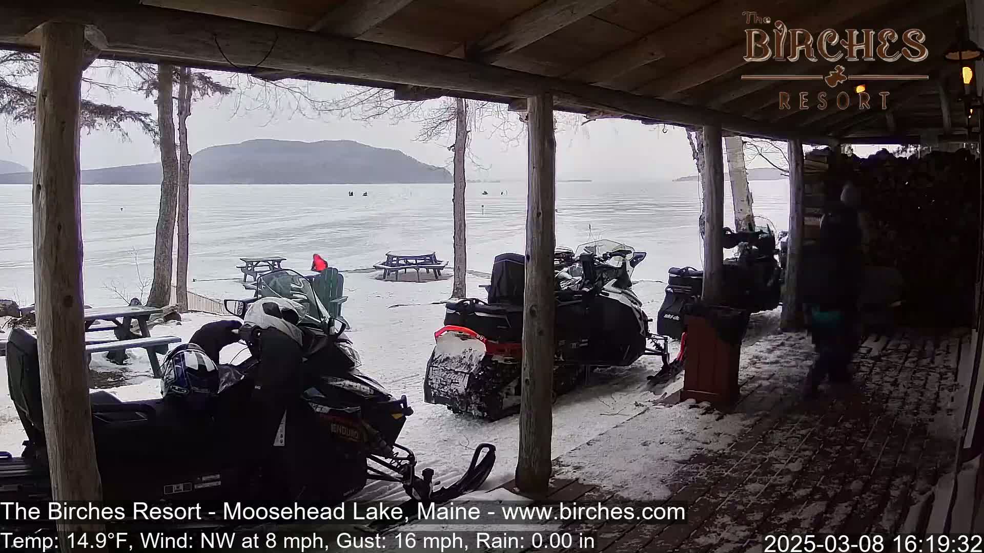 Several snowmobiles are parked under a wooden porch overlooking a snow-covered lake and distant mountains under an overcast sky.