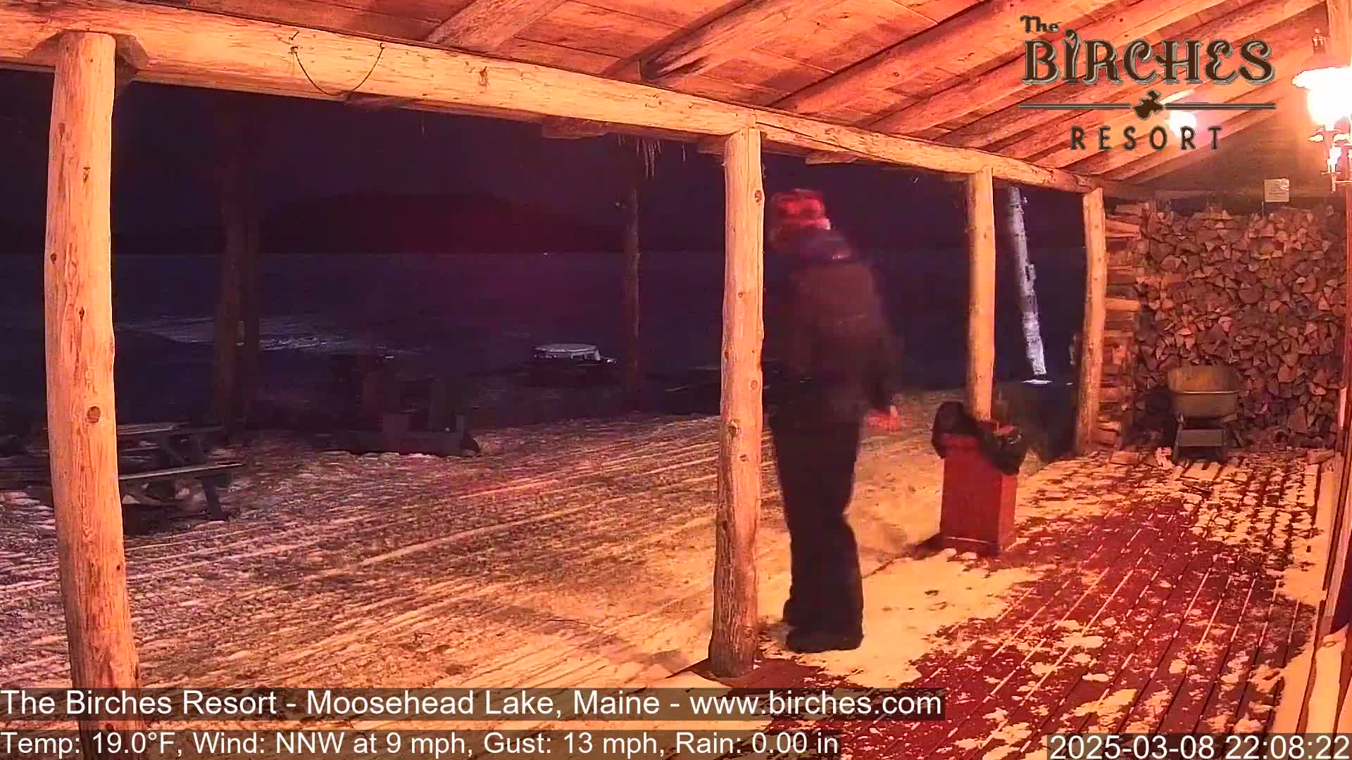 A person walks across a snow-covered porch of a wooden building at night during a light snowfall with a northwest wind.