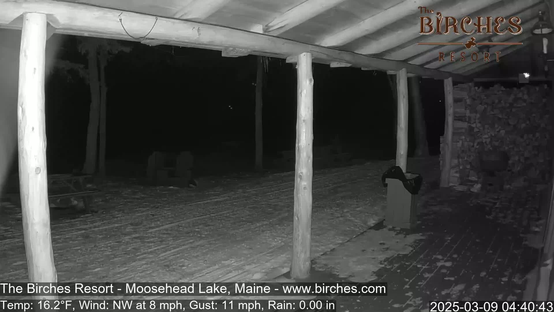 A nighttime view of a snow-covered porch with wooden posts and a woodpile, under a northwest wind of 8 mph with gusts up to 11 mph and a temperature of 16.2°F.