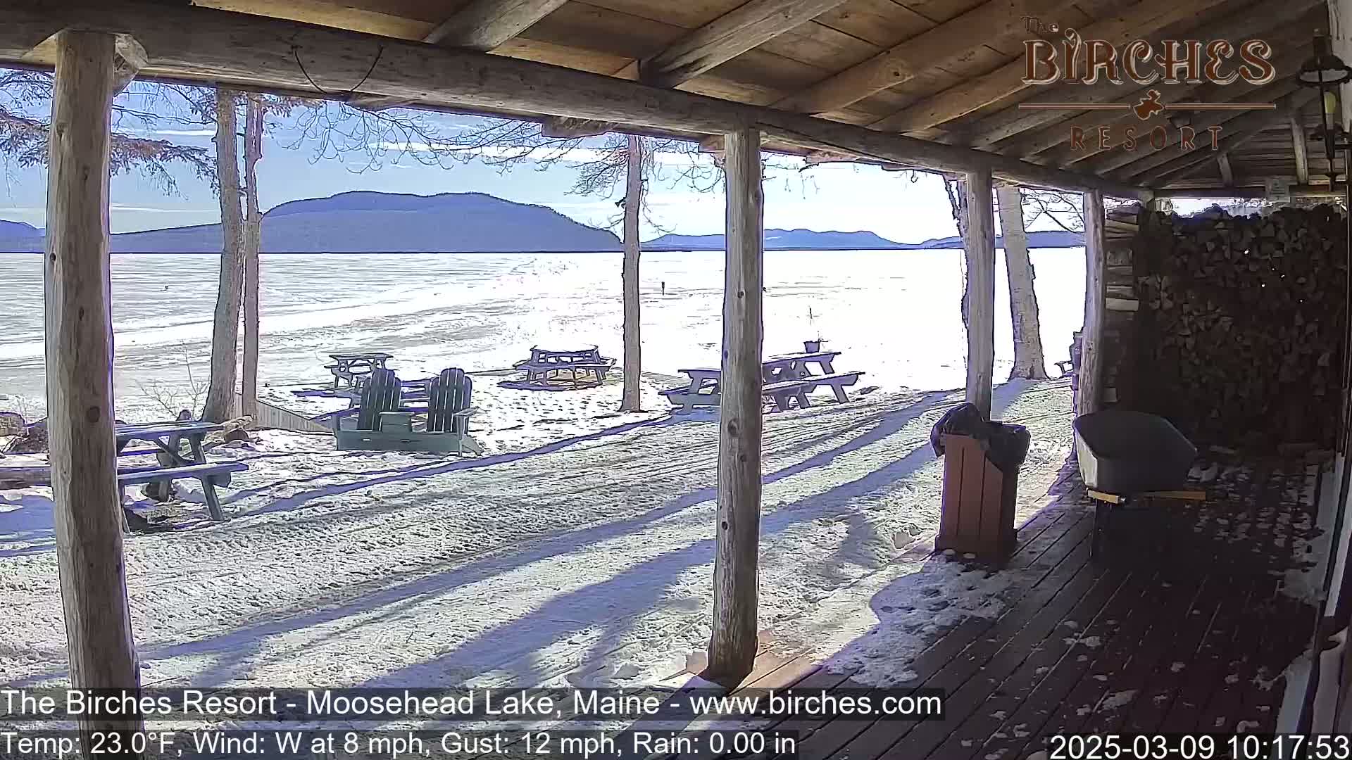 A partly sunny view of a snow-covered lakeshore is seen from a wooden porch, with picnic tables and Adirondack chairs scattered across the snow.