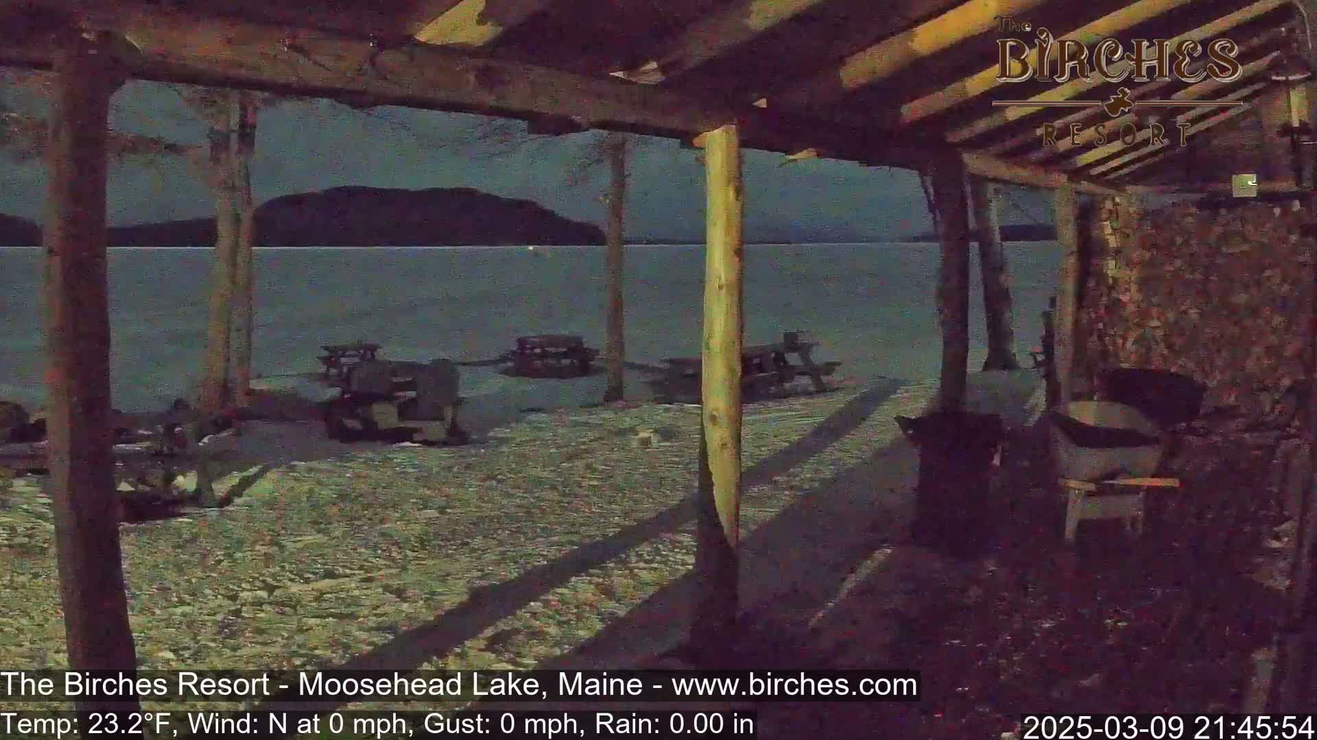 A snow-covered lakeside patio with picnic tables is visible under a nighttime sky from within a covered structure.