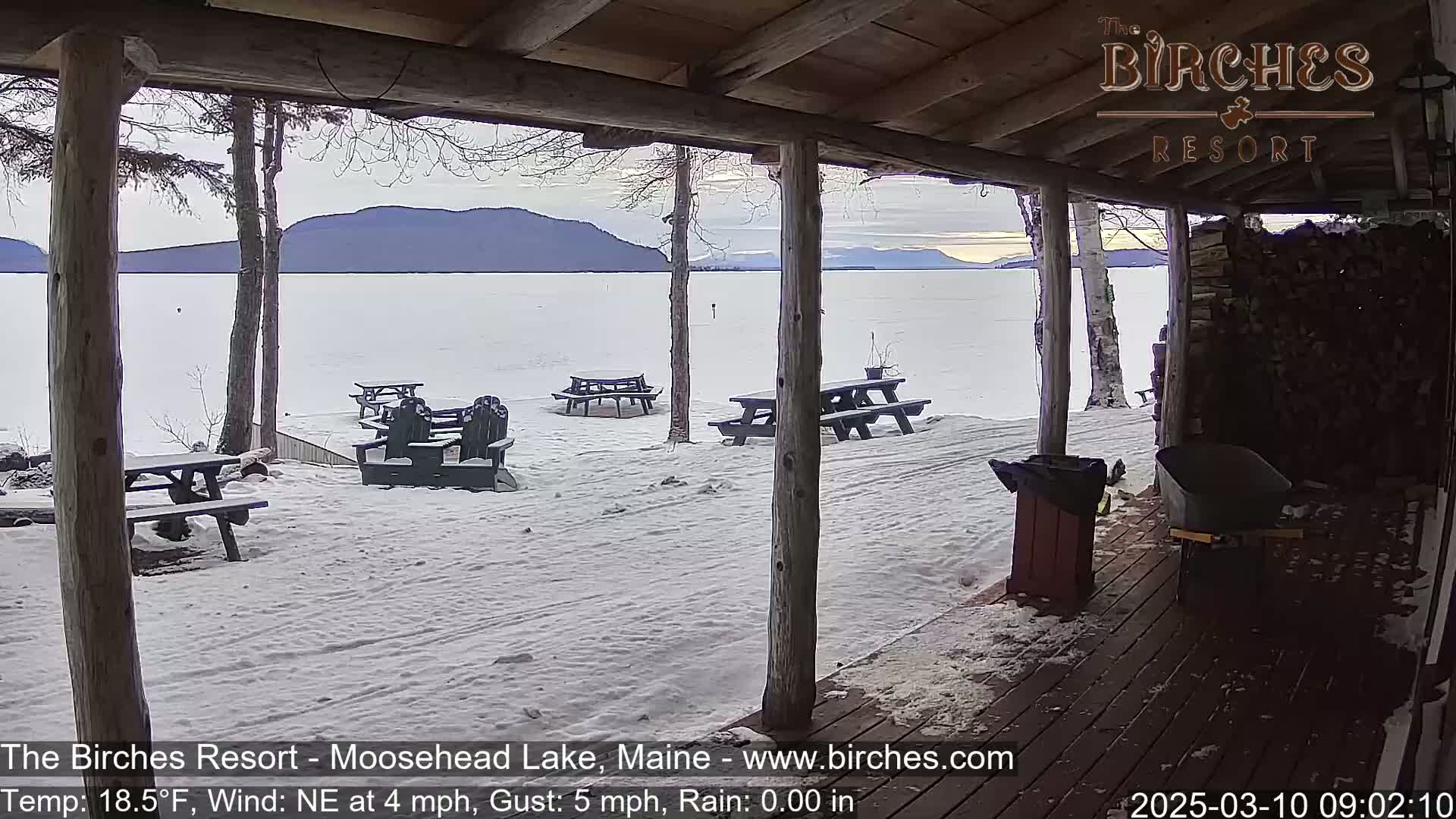 A snow-covered lakeside patio with picnic tables and chairs is seen from a covered porch, with mountains in the background under a partly cloudy sky.