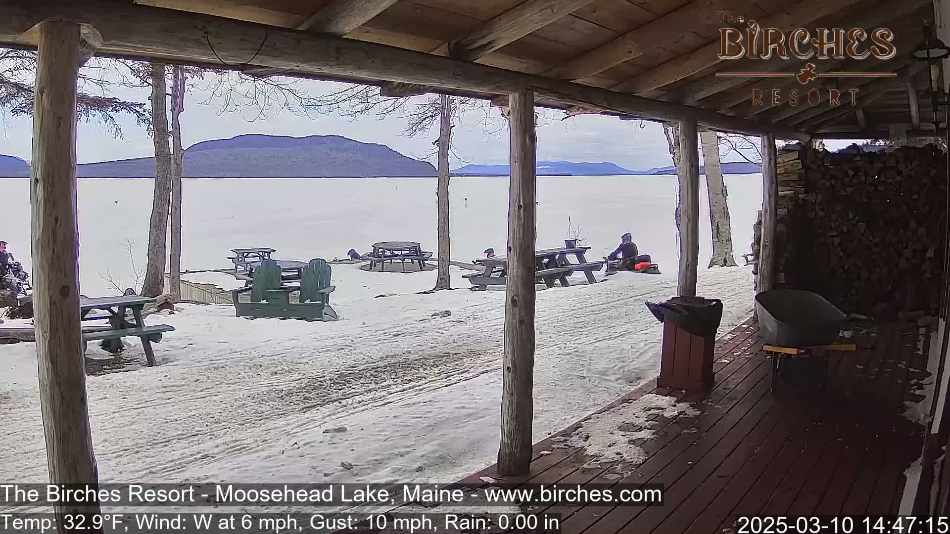 A snow-covered lakeside patio with picnic tables and a person on a snowmobile is seen from a porch on a partly cloudy day with a light wind.