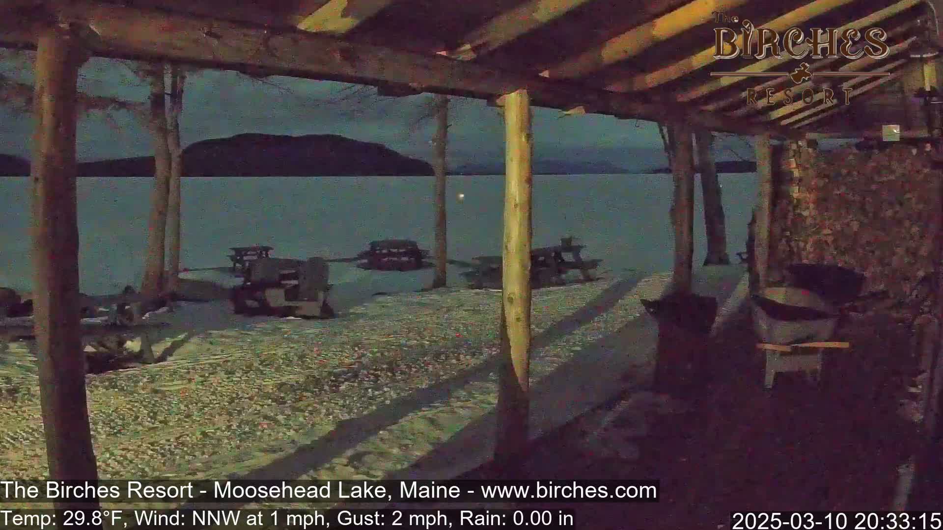 A snow-covered lakeside area at night is viewed from under a wooden shelter, with picnic tables scattered nearby and a calm lake visible in the background.
