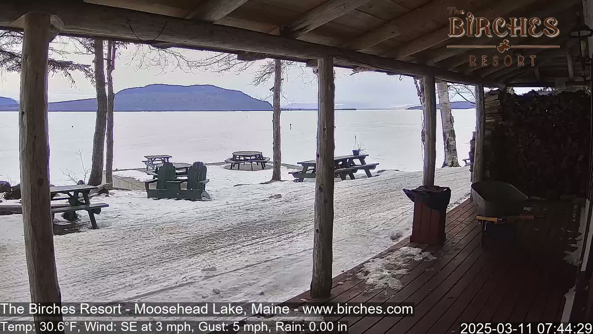 A snow-covered lakeside patio with picnic tables and Adirondack chairs is viewed from a wooden porch, overlooking a partially frozen lake and distant mountains under a cloudy sky.