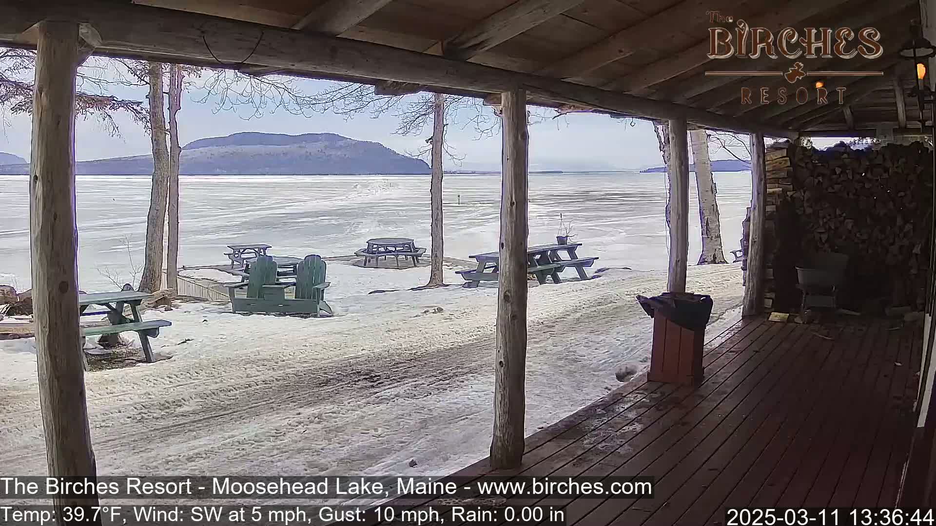 A partially frozen lake is visible from a wooden porch, with several picnic tables and chairs scattered across the snow-covered ground in the background; it's a partly sunny, windy day.