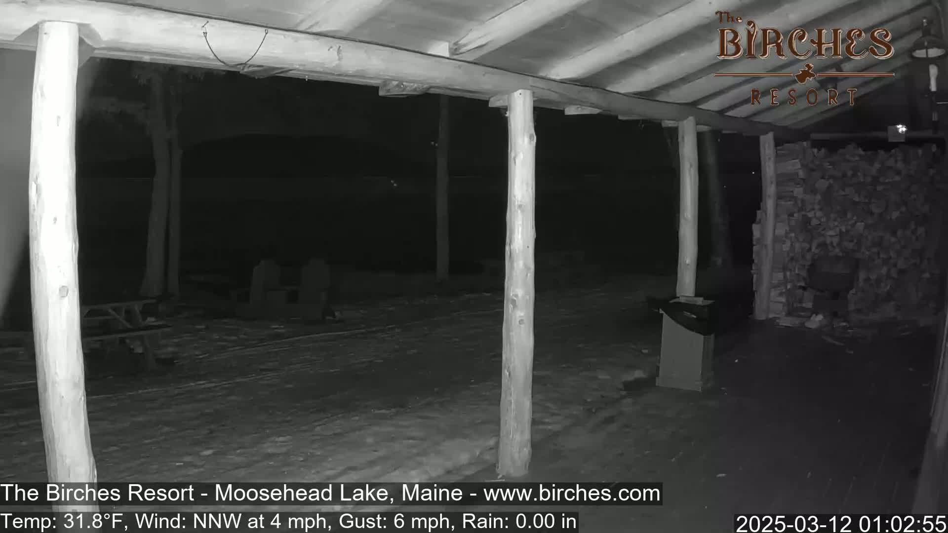 A nighttime, snowy scene under a covered porch shows wooden posts, picnic tables, and a stack of firewood with a 4 mph wind and a temperature of 31.8°F.