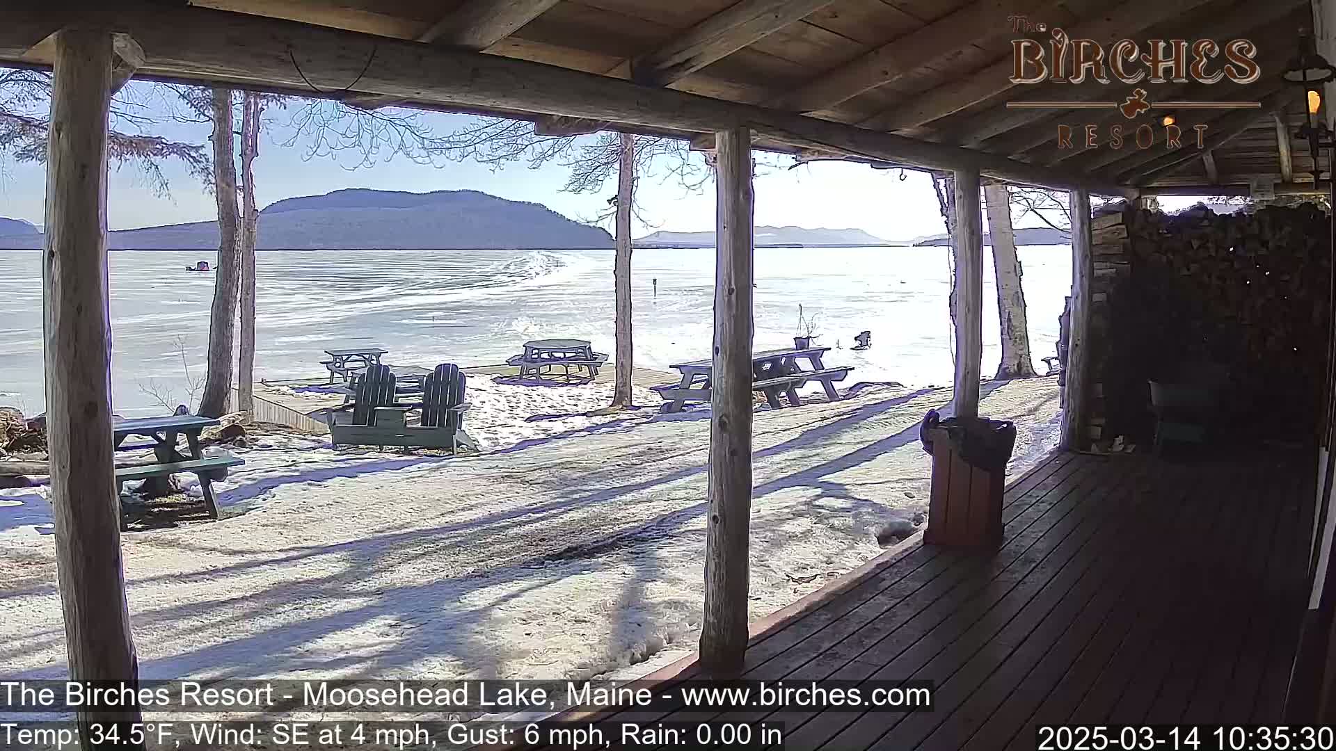 A partly sunny view of a partially frozen lake from a wooden porch with picnic tables and Adirondack chairs, some snow on the ground.