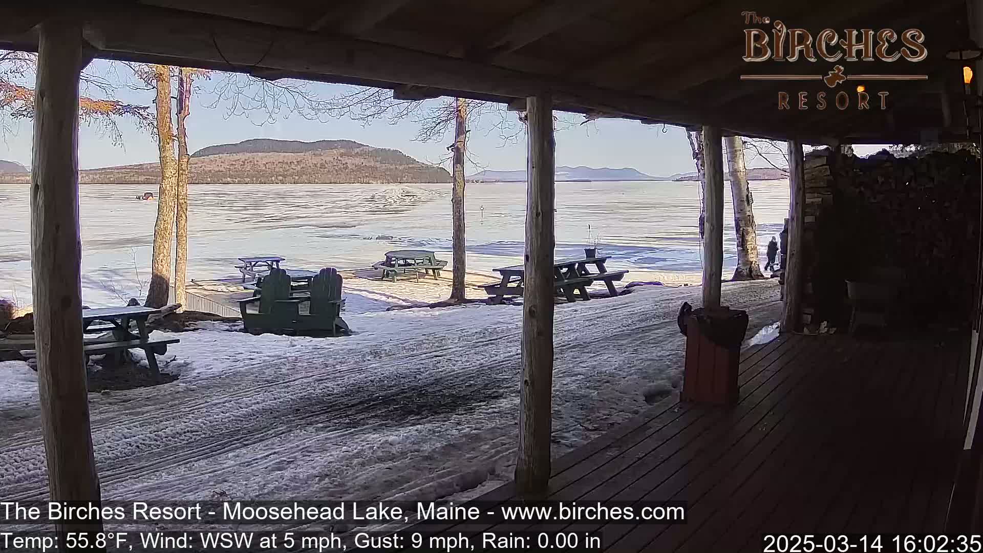 A partly sunny view of a partially frozen lake with snow-covered ground and picnic tables seen from a wooden porch.