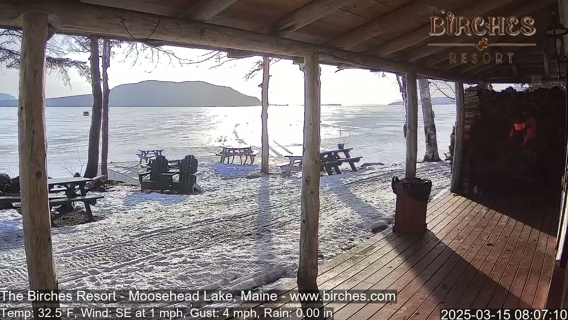 A snow-covered lakeside patio with picnic tables and chairs is partially visible from inside a wooden porch, overlooking a partly frozen lake and distant mountains under a partly sunny sky.