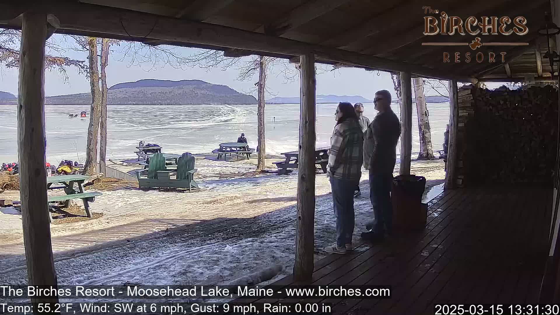 From a covered porch, three people stand looking out at a partially frozen lake with snow on the ground under a sunny sky, and mountains in the distance.