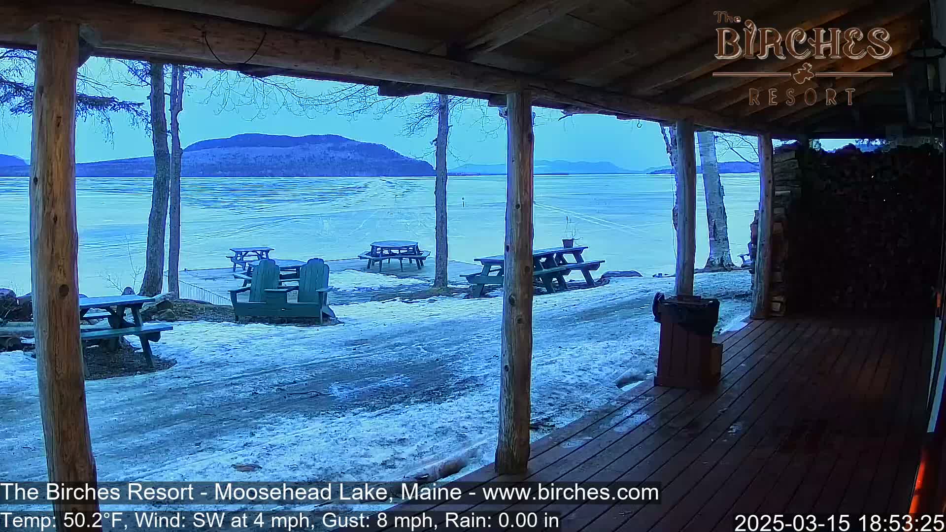A partially frozen lake is viewed from a wooden porch with picnic tables and chairs scattered on the snow-covered ground; the background shows distant mountains under a clear, twilight sky.