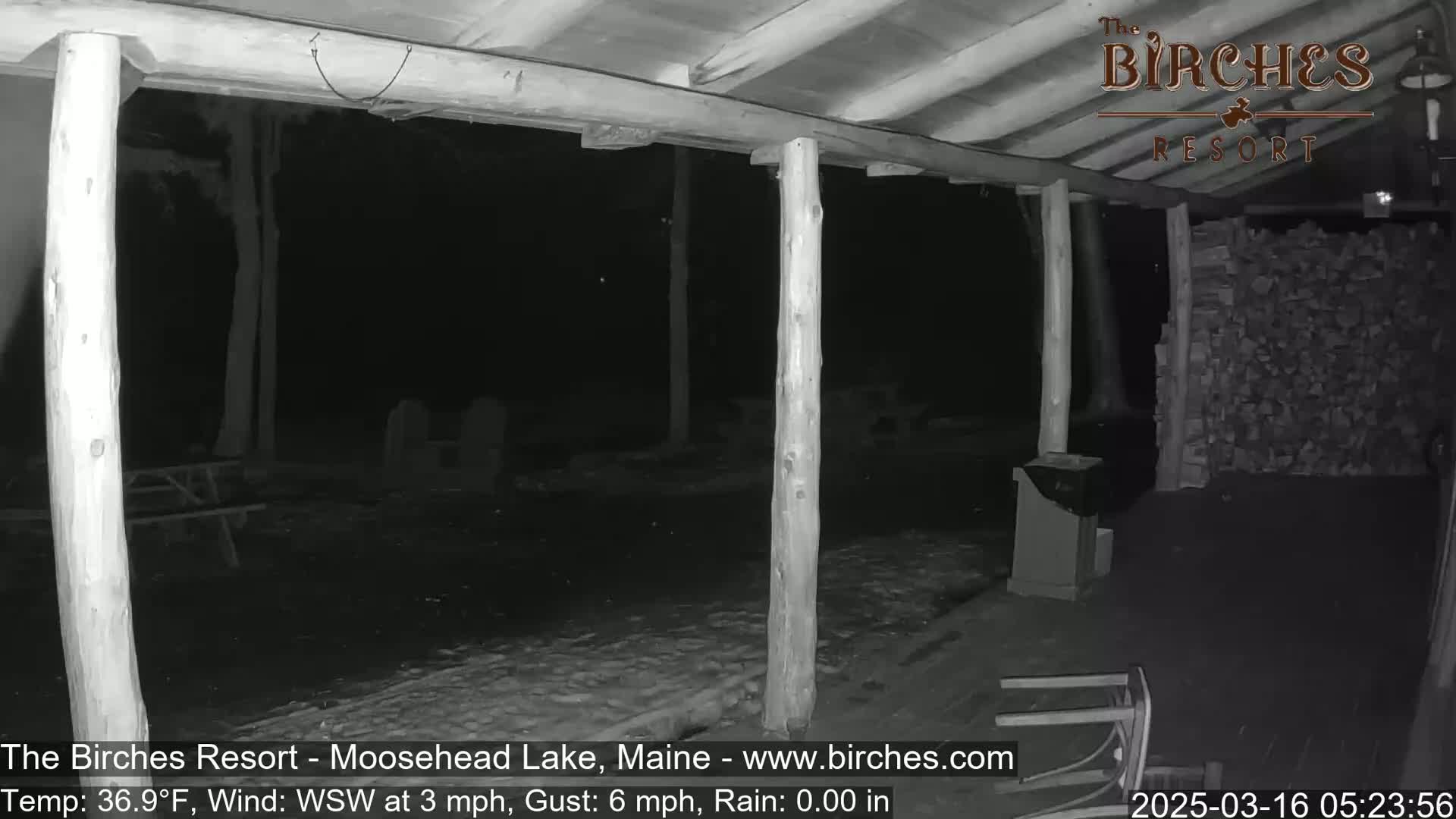 A nighttime view from a covered porch shows a snow-covered ground, picnic tables, and a woodpile, with a wind from the west-southwest at 3 mph and gusts up to 6 mph.