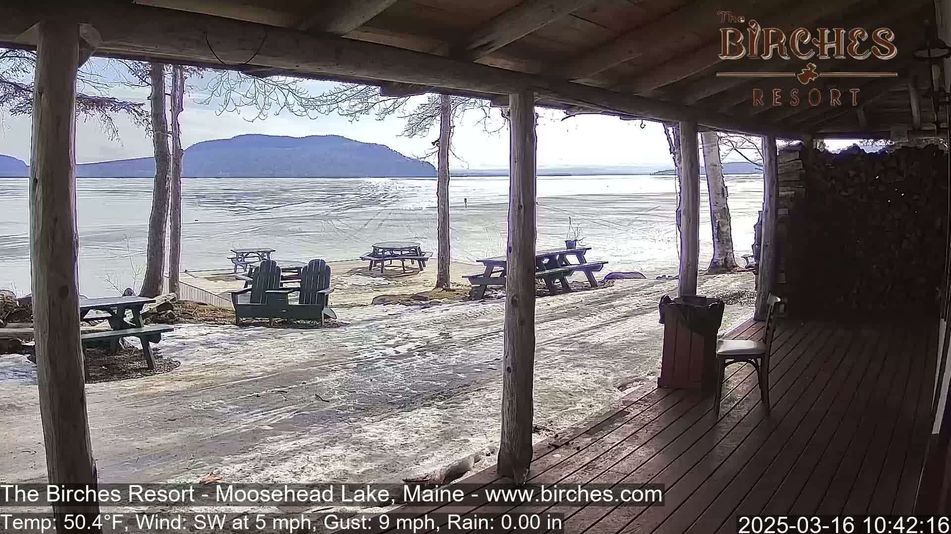 A partially covered porch overlooks a partially frozen lake with mountains in the background on a partly sunny day with a southwest wind.