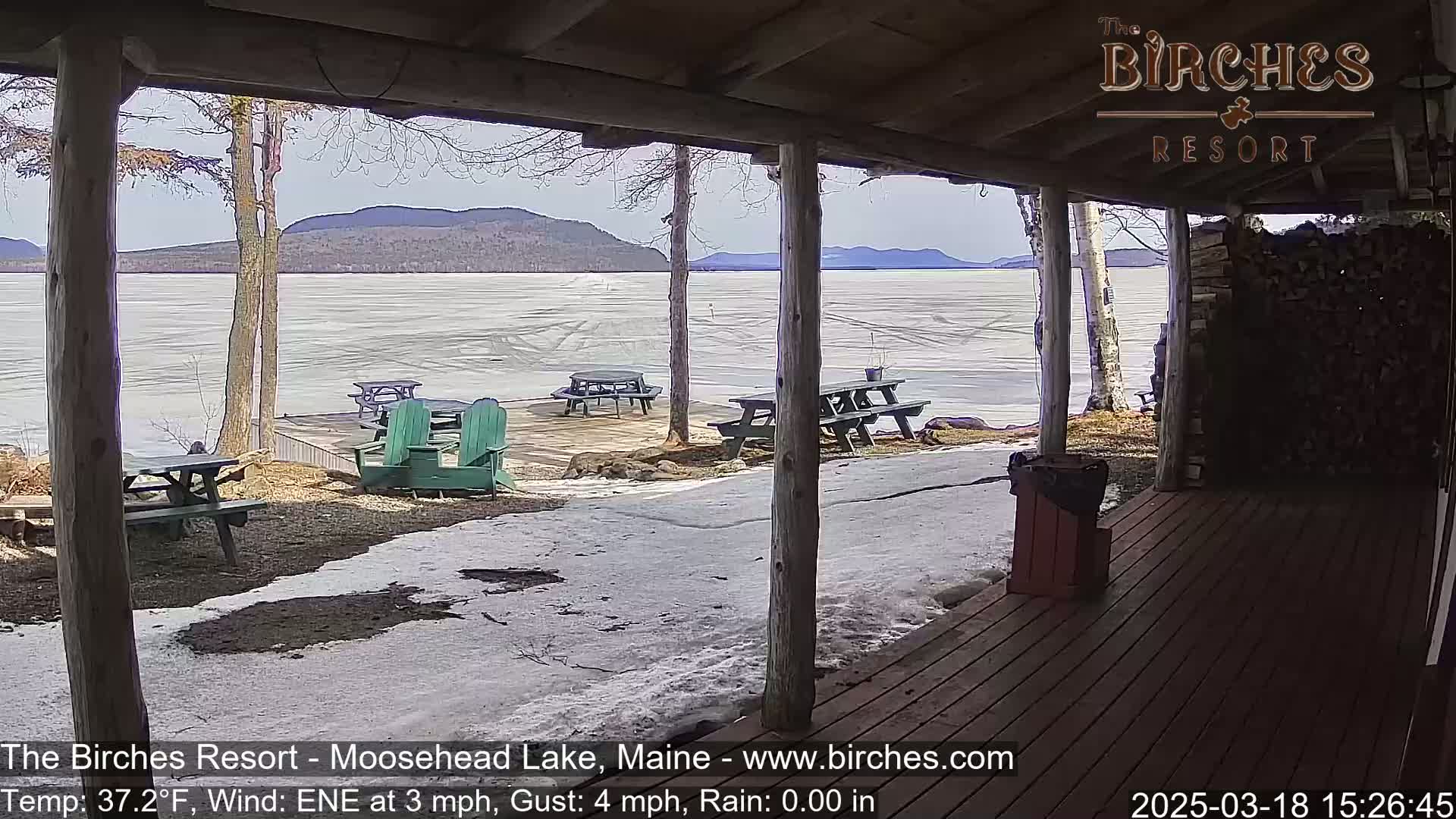 A partially frozen lake is visible from a covered porch with picnic tables and Adirondack chairs, under partly cloudy skies.