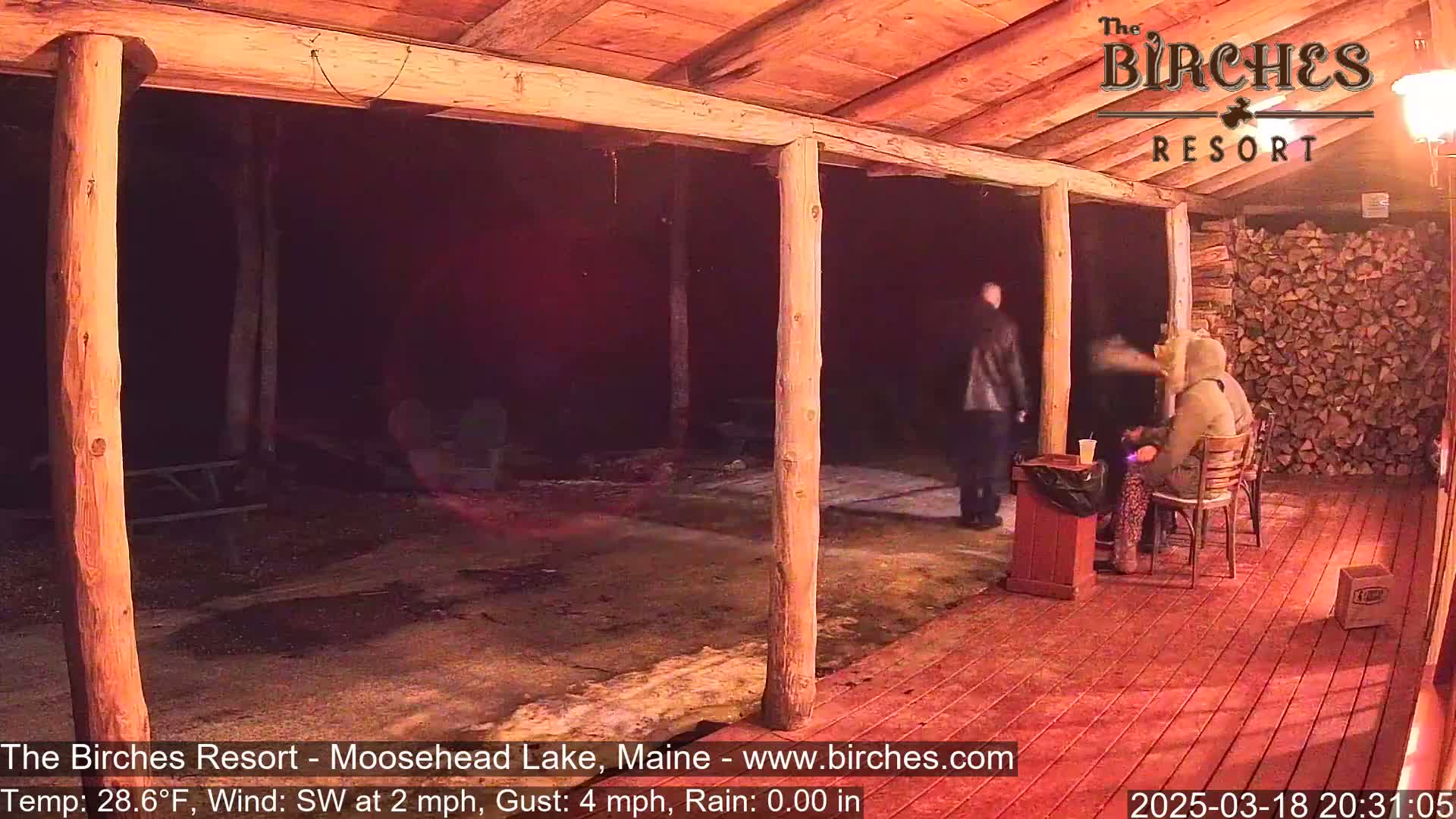A dimly lit, wooden-post covered porch shows two people sitting at a table, one person standing, and a pile of firewood to the right, under a night sky with a light rain and a temperature of 28.6°F.