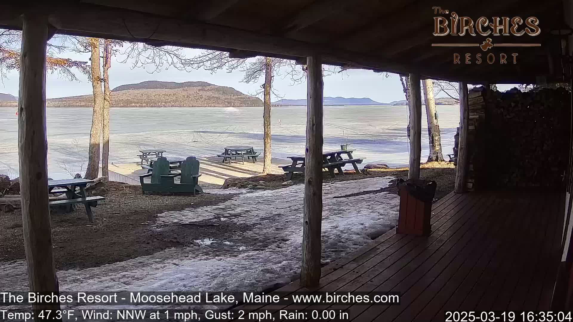 A partly sunny view of a frozen lake with picnic tables and Adirondack chairs on a snow-dusted patio seen from a covered porch.