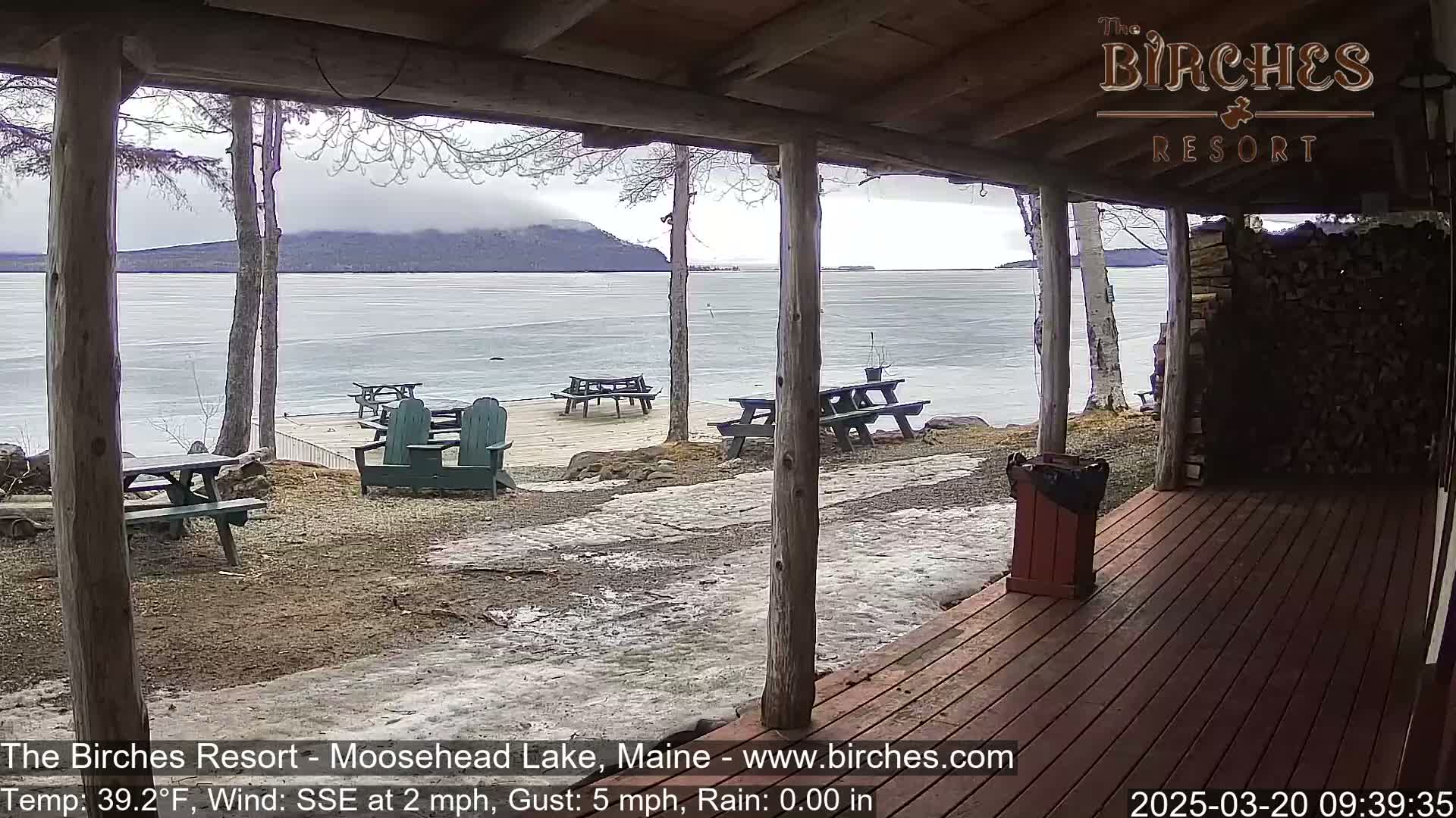 A partially covered wooden deck overlooks a partially frozen lake under an overcast sky, with several picnic tables and Adirondack chairs scattered on the ground.