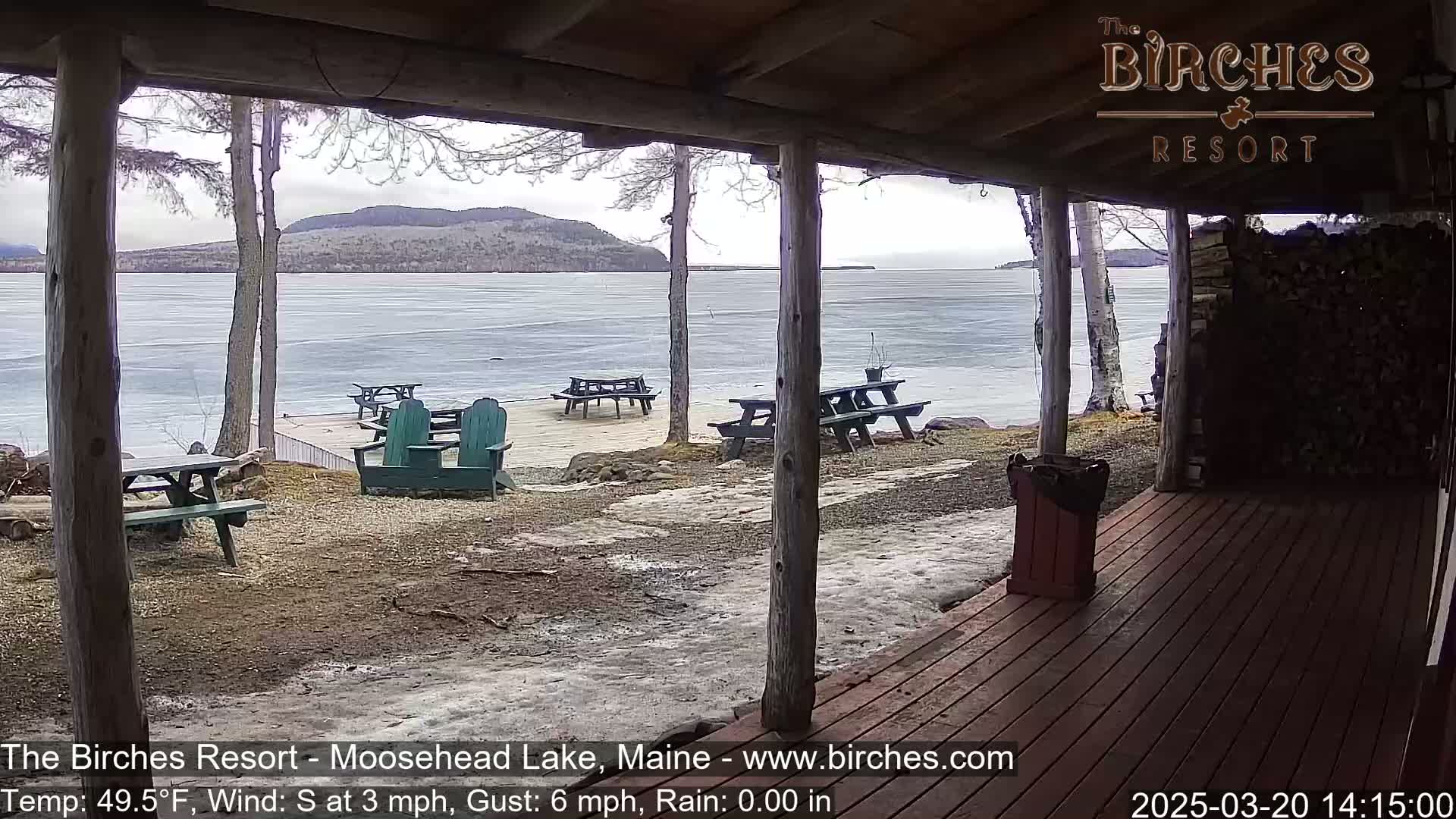 From a covered porch, a partially frozen lake is visible under an overcast sky, with several picnic tables and Adirondack chairs situated on the lakeshore.