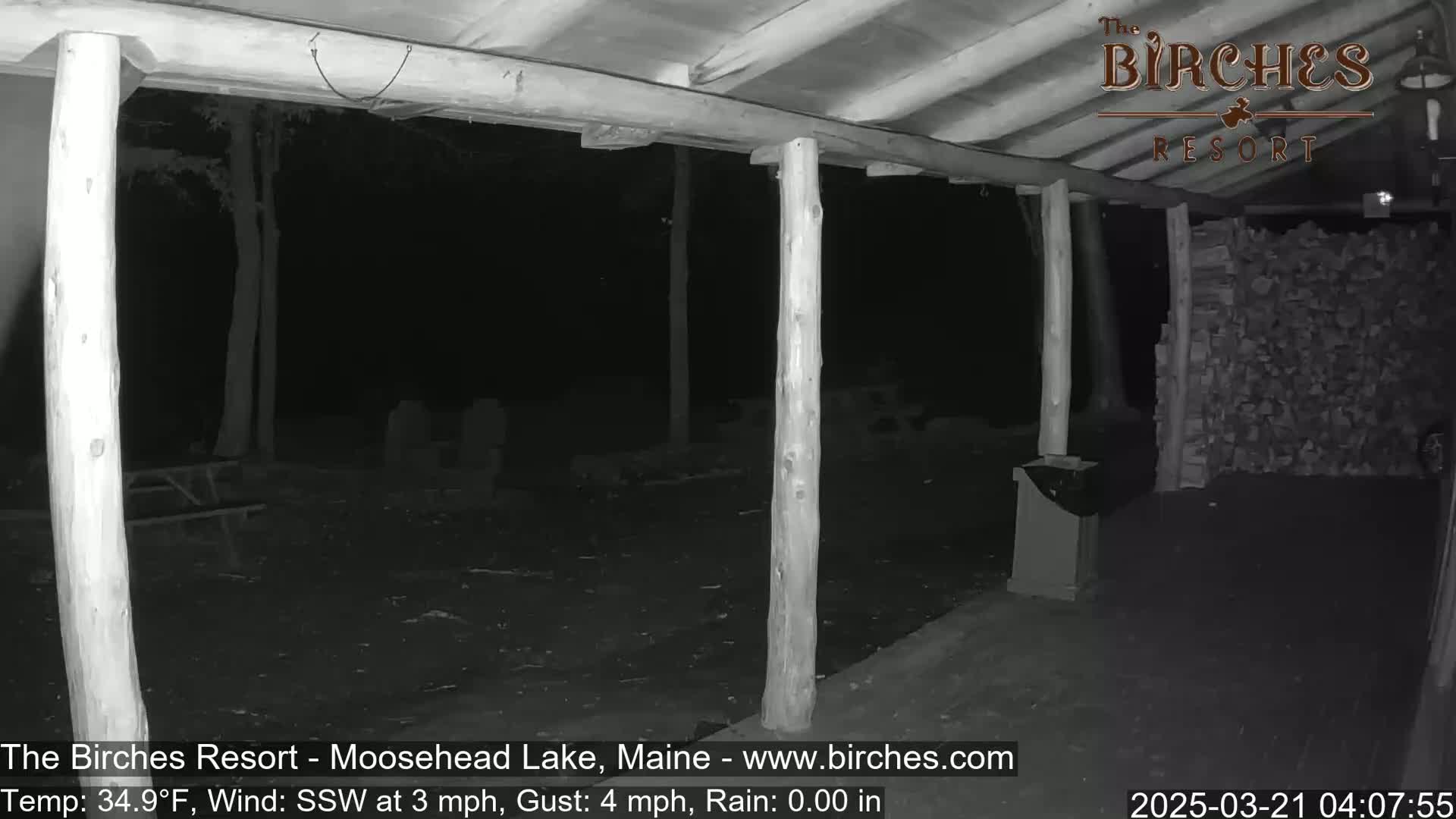 A nighttime, rainy view from under a covered porch shows a wood-stacked wall, picnic tables, and surrounding trees.