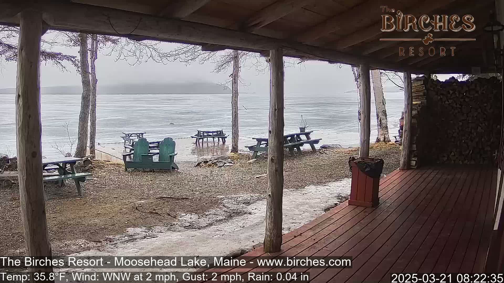 A view from a covered porch shows a partly icy lake under a gray, rainy sky, with several picnic tables and Adirondack chairs on the lakeshore.