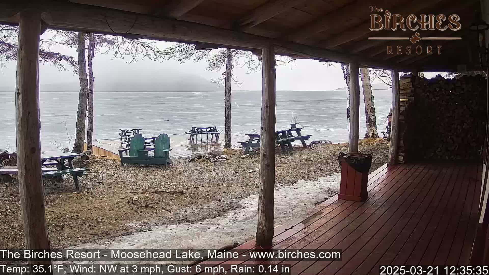 From a covered porch, several picnic tables and Adirondack chairs sit on a gravel area beside a rain-swept lake partially covered in ice.