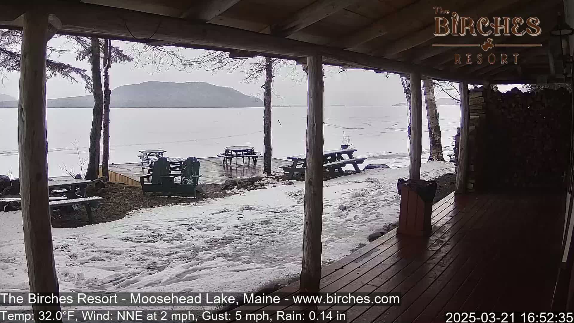 A partially covered wooden deck overlooks a partially frozen lake under an overcast sky, with snow on the ground and picnic tables visible.