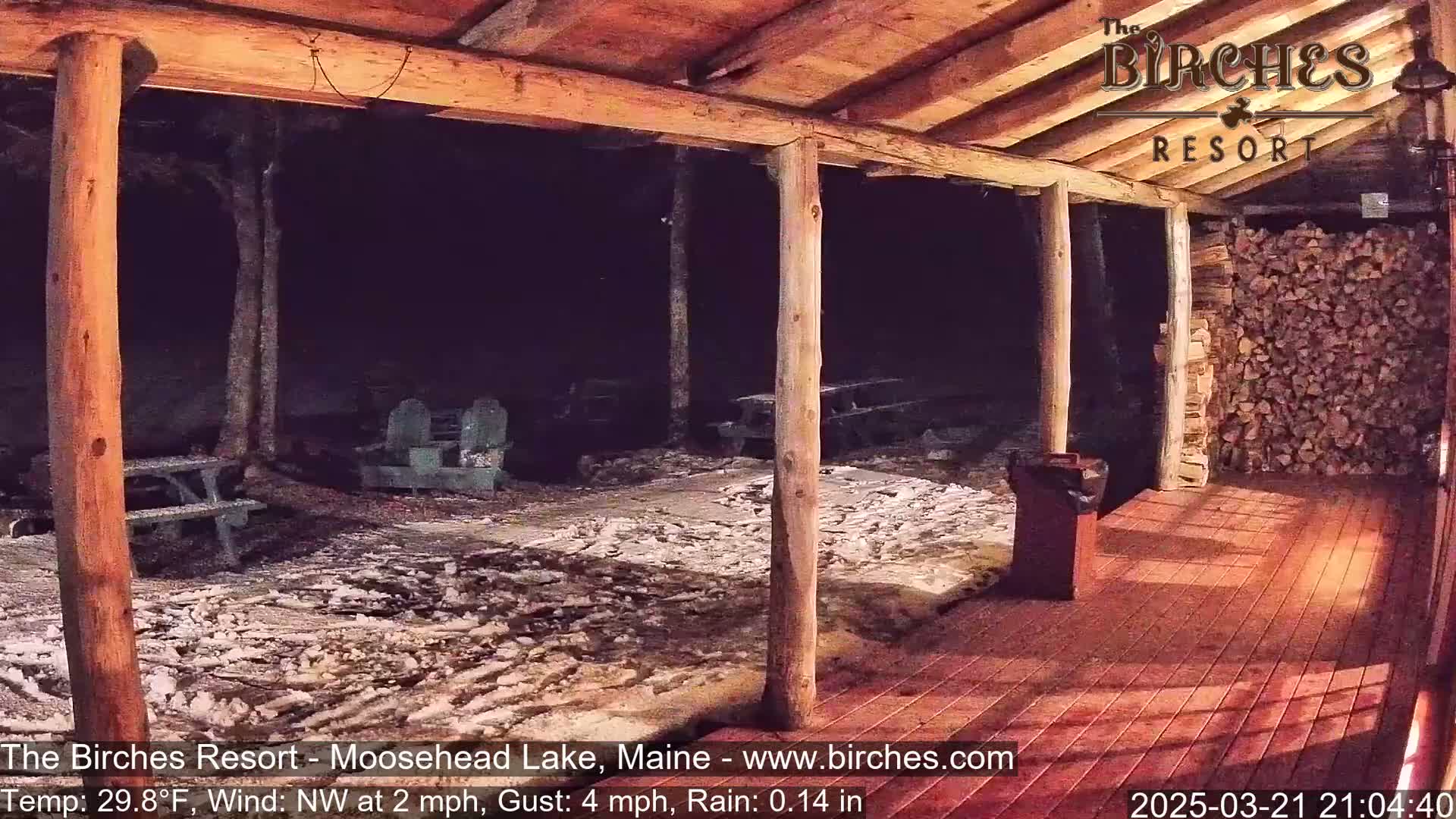 A nighttime view from a wooden porch shows a snow-dusted ground with picnic tables and chairs under a dark sky with light rain.