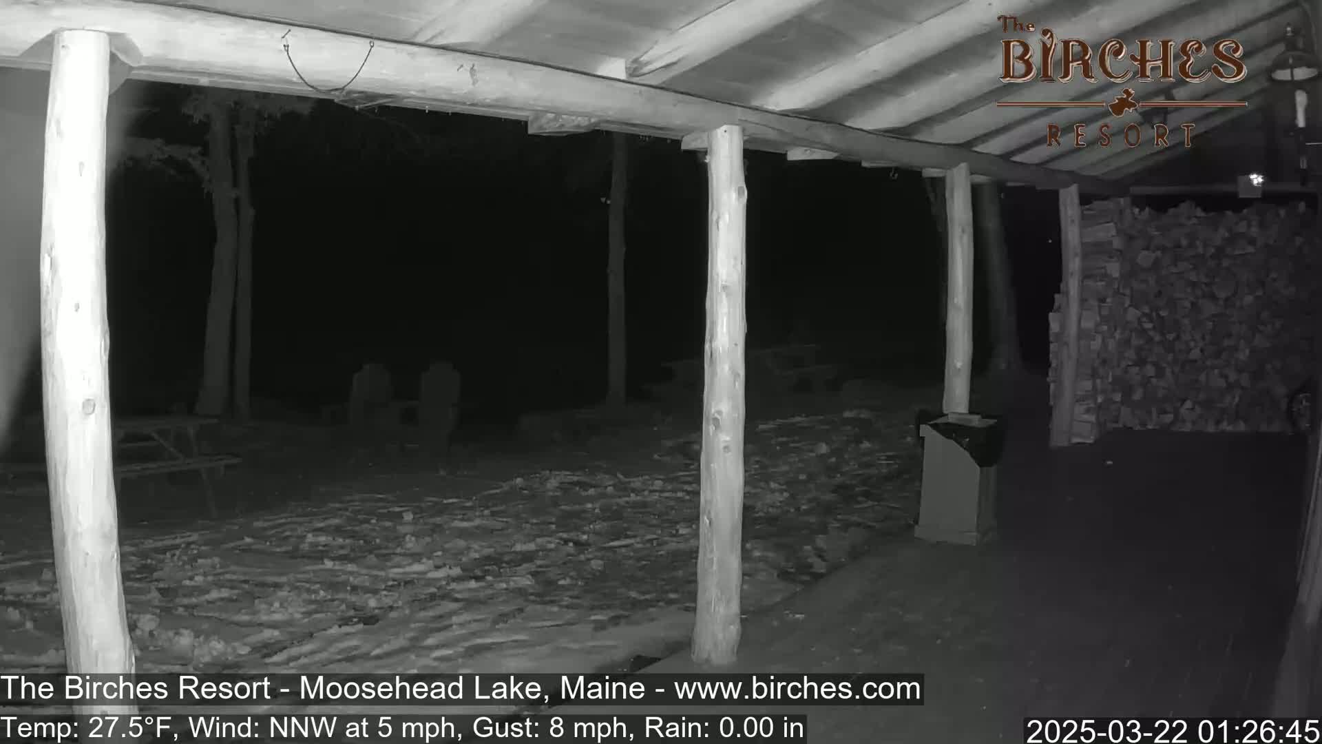 A snow-covered porch is seen at night under a covered wooden structure, with a woodpile visible to the right.