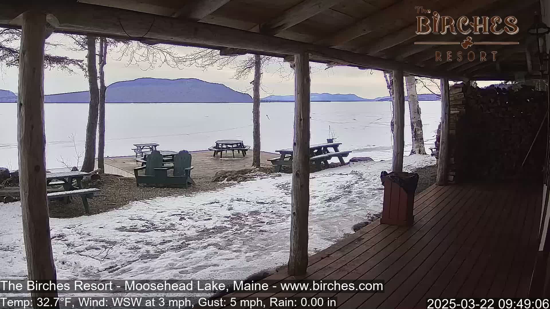 A partially frozen lake is viewed from a covered porch, with several picnic tables and chairs on the snow-covered ground, under a mostly cloudy sky.