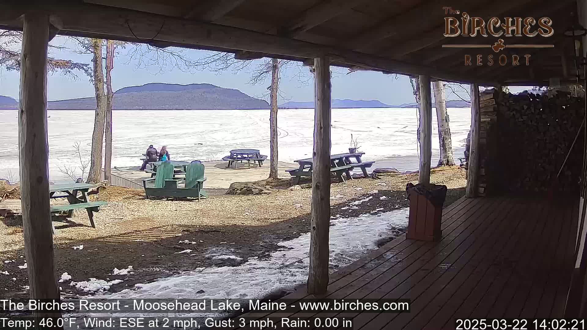 From a covered porch, a partially frozen lake is visible under a sunny sky, with several picnic tables and a few people sitting outside.