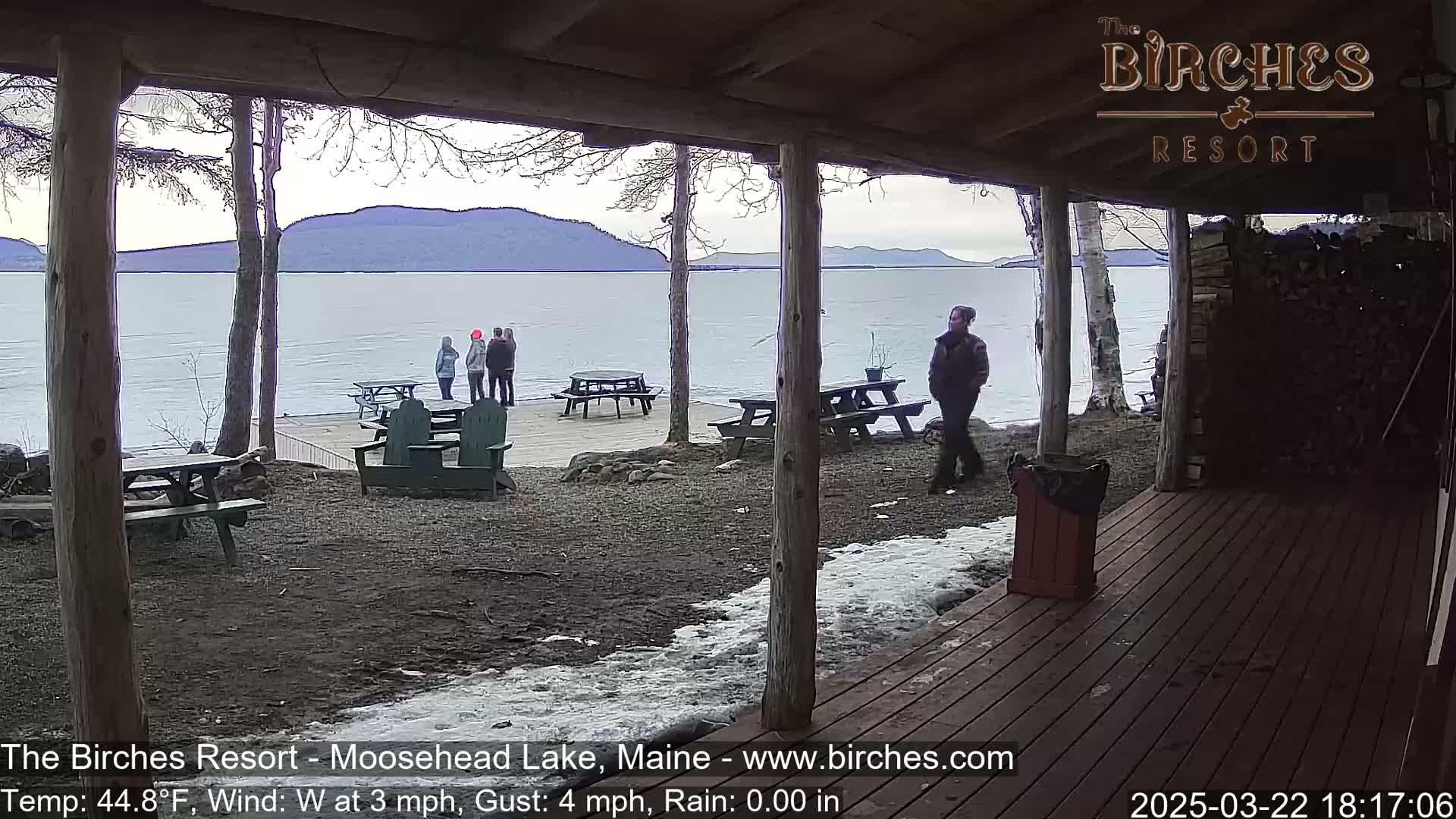 A lakeside patio with picnic tables and Adirondack chairs overlooks a calm lake and distant mountains under an overcast sky.