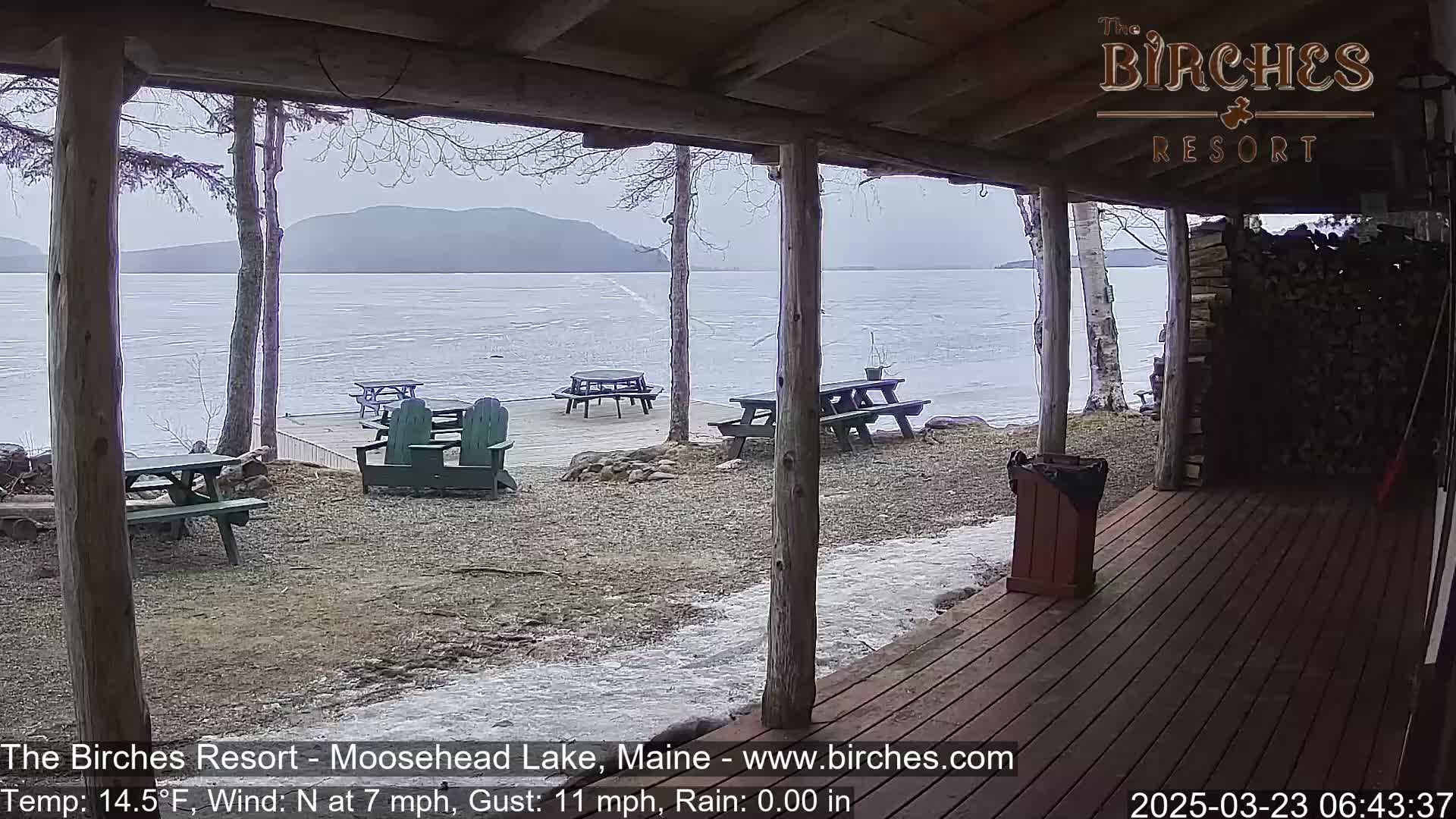 A partially frozen lake is viewed from a wooden porch with picnic tables and Adirondack chairs, under an overcast sky.