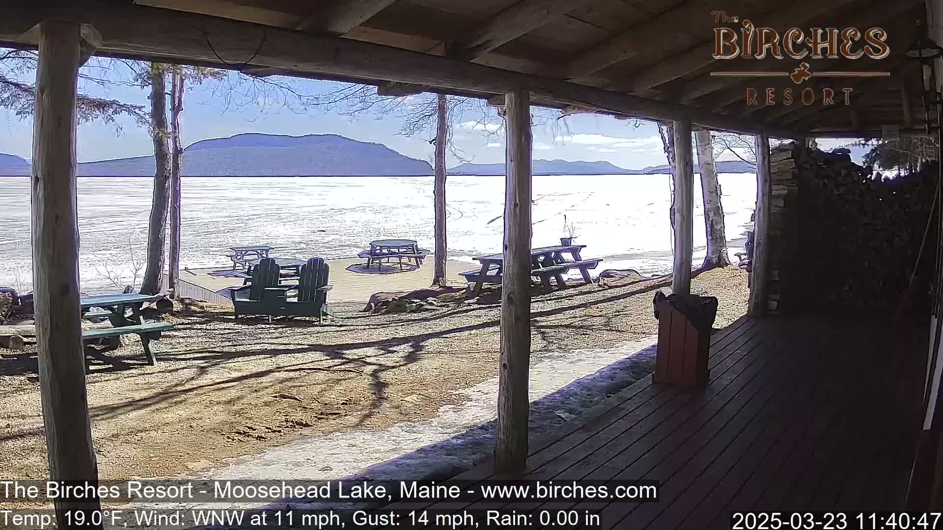 A partly sunny view from a porch shows a partially frozen lake with picnic tables and Adirondack chairs on the lakeshore under a mostly clear sky with a light wind.