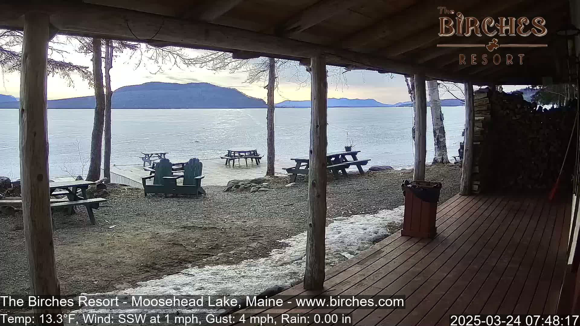 A partially covered porch overlooks a partly frozen lake with mountains in the distance, under a cloudy sky.